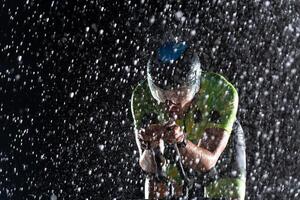 A triathlete braving the rain as he cycles through the night, preparing himself for the upcoming marathon. The blurred raindrops in the foreground and the dark, moody atmosphere in the background add photo