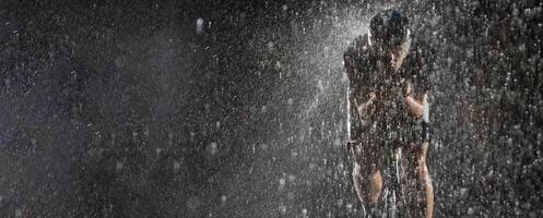 A triathlete braving the rain as he cycles through the night, preparing himself for the upcoming marathon. The blurred raindrops in the foreground and the dark, moody atmosphere in the background add photo