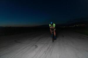A triathlete rides his bike in the darkness of night, pushing himself to prepare for a marathon. The contrast between the darkness and the light of his bike creates a sense of drama and highlights the photo