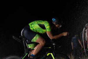 A triathlete braving the rain as he cycles through the night, preparing himself for the upcoming marathon. The blurred raindrops in the foreground and the dark, moody atmosphere in the background add photo