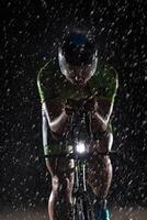 A triathlete braving the rain as he cycles through the night, preparing himself for the upcoming marathon. The blurred raindrops in the foreground and the dark, moody atmosphere in the background add photo