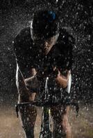 A triathlete braving the rain as he cycles through the night, preparing himself for the upcoming marathon. The blurred raindrops in the foreground and the dark, moody atmosphere in the background add photo