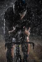 A triathlete braving the rain as he cycles through the night, preparing himself for the upcoming marathon. The blurred raindrops in the foreground and the dark, moody atmosphere in the background add photo