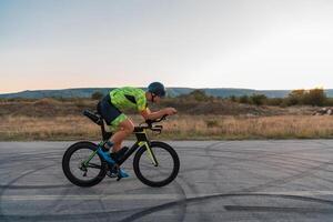 Triathlete riding his bicycle during sunset, preparing for a marathon. The warm colors of the sky provide a beautiful backdrop for his determined and focused effort. photo