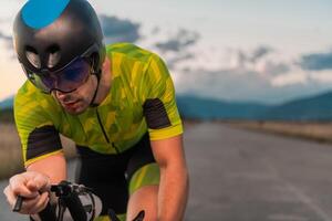 Close up photo of triathlete riding his bicycle during sunset, preparing for a marathon. The warm colors of the sky provide a beautiful backdrop for his determined and focused effort.