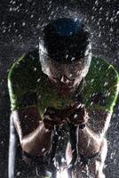 A triathlete braving the rain as he cycles through the night, preparing himself for the upcoming marathon. The blurred raindrops in the foreground and the dark, moody atmosphere in the background add photo