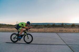 Triathlete riding his bicycle during sunset, preparing for a marathon. The warm colors of the sky provide a beautiful backdrop for his determined and focused effort. photo