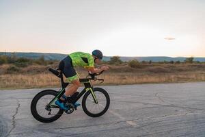 Triathlete riding his bicycle during sunset, preparing for a marathon. The warm colors of the sky provide a beautiful backdrop for his determined and focused effort. photo