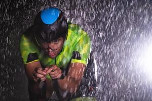 A triathlete braving the rain as he cycles through the night, preparing himself for the upcoming marathon. The blurred raindrops in the foreground and the dark, moody atmosphere in the background add photo