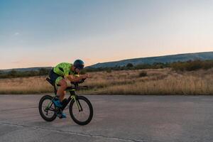 Triathlete riding his bicycle during sunset, preparing for a marathon. The warm colors of the sky provide a beautiful backdrop for his determined and focused effort. photo