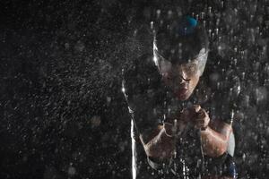 A triathlete braving the rain as he cycles through the night, preparing himself for the upcoming marathon. The blurred raindrops in the foreground and the dark, moody atmosphere in the background add photo