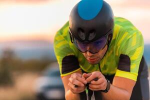 Close up photo of triathlete riding his bicycle during sunset, preparing for a marathon. The warm colors of the sky provide a beautiful backdrop for his determined and focused effort.
