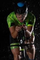 A triathlete braving the rain as he cycles through the night, preparing himself for the upcoming marathon. The blurred raindrops in the foreground and the dark, moody atmosphere in the background add photo