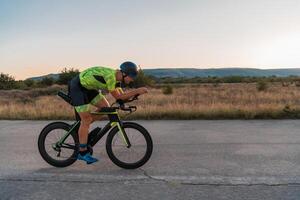 Triathlete riding his bicycle during sunset, preparing for a marathon. The warm colors of the sky provide a beautiful backdrop for his determined and focused effort. photo
