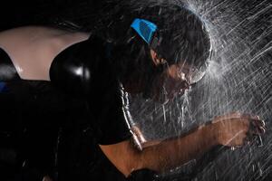 A triathlete braving the rain as he cycles through the night, preparing himself for the upcoming marathon. The blurred raindrops in the foreground and the dark, moody atmosphere in the background add photo