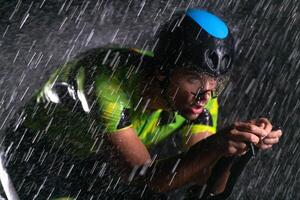 A triathlete braving the rain as he cycles through the night, preparing himself for the upcoming marathon. The blurred raindrops in the foreground and the dark, moody atmosphere in the background add photo