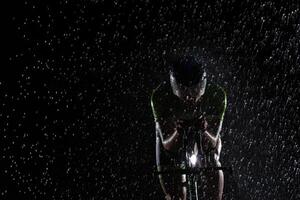 A triathlete braving the rain as he cycles through the night, preparing himself for the upcoming marathon. The blurred raindrops in the foreground and the dark, moody atmosphere in the background add photo