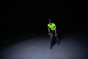 A triathlete rides his bike in the darkness of night, pushing himself to prepare for a marathon. The contrast between the darkness and the light of his bike creates a sense of drama and highlights the photo