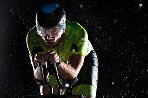 A triathlete braving the rain as he cycles through the night, preparing himself for the upcoming marathon. The blurred raindrops in the foreground and the dark, moody atmosphere in the background add photo