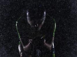 A triathlete braving the rain as he cycles through the night, preparing himself for the upcoming marathon. The blurred raindrops in the foreground and the dark, moody atmosphere in the background add photo