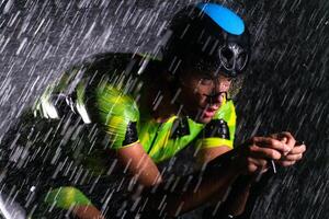 A triathlete braving the rain as he cycles through the night, preparing himself for the upcoming marathon. The blurred raindrops in the foreground and the dark, moody atmosphere in the background add photo