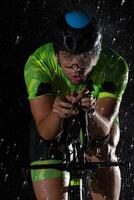 A triathlete braving the rain as he cycles through the night, preparing himself for the upcoming marathon. The blurred raindrops in the foreground and the dark, moody atmosphere in the background add photo
