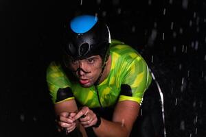 A triathlete braving the rain as he cycles through the night, preparing himself for the upcoming marathon. The blurred raindrops in the foreground and the dark, moody atmosphere in the background add photo