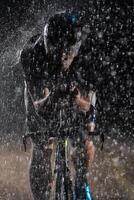 A triathlete braving the rain as he cycles through the night, preparing himself for the upcoming marathon. The blurred raindrops in the foreground and the dark, moody atmosphere in the background add photo