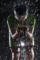 A triathlete braving the rain as he cycles through the night, preparing himself for the upcoming marathon. The blurred raindrops in the foreground and the dark, moody atmosphere in the background add photo