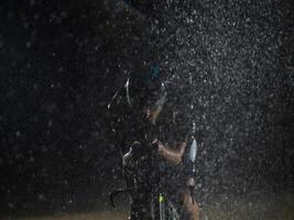 A triathlete braving the rain as he cycles through the night, preparing himself for the upcoming marathon. The blurred raindrops in the foreground and the dark, moody atmosphere in the background add photo