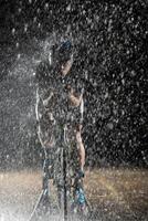 A triathlete braving the rain as he cycles through the night, preparing himself for the upcoming marathon. The blurred raindrops in the foreground and the dark, moody atmosphere in the background add photo