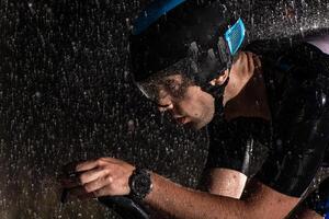 A triathlete braving the rain as he cycles through the night, preparing himself for the upcoming marathon. The blurred raindrops in the foreground and the dark, moody atmosphere in the background add photo