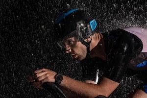 A triathlete braving the rain as he cycles through the night, preparing himself for the upcoming marathon. The blurred raindrops in the foreground and the dark, moody atmosphere in the background add photo