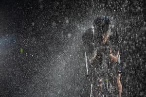 A triathlete braving the rain as he cycles through the night, preparing himself for the upcoming marathon. The blurred raindrops in the foreground and the dark, moody atmosphere in the background add photo