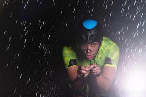 A triathlete braving the rain as he cycles through the night, preparing himself for the upcoming marathon. The blurred raindrops in the foreground and the dark, moody atmosphere in the background add photo