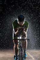 A triathlete braving the rain as he cycles through the night, preparing himself for the upcoming marathon. The blurred raindrops in the foreground and the dark, moody atmosphere in the background add photo