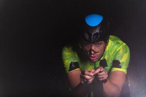 A triathlete braving the rain as he cycles through the night, preparing himself for the upcoming marathon. The blurred raindrops in the foreground and the dark, moody atmosphere in the background add photo