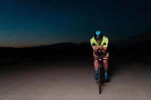 A triathlete rides his bike in the darkness of night, pushing himself to prepare for a marathon. The contrast between the darkness and the light of his bike creates a sense of drama and highlights the photo
