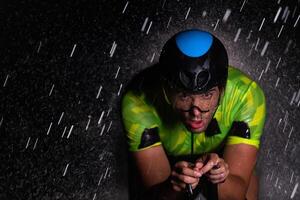 A triathlete braving the rain as he cycles through the night, preparing himself for the upcoming marathon. The blurred raindrops in the foreground and the dark, moody atmosphere in the background add photo