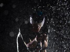 A triathlete braving the rain as he cycles through the night, preparing himself for the upcoming marathon. The blurred raindrops in the foreground and the dark, moody atmosphere in the background add photo