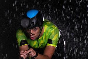 A triathlete braving the rain as he cycles through the night, preparing himself for the upcoming marathon. The blurred raindrops in the foreground and the dark, moody atmosphere in the background add photo