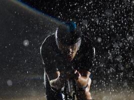 A triathlete braving the rain as he cycles through the night, preparing himself for the upcoming marathon. The blurred raindrops in the foreground and the dark, moody atmosphere in the background add photo