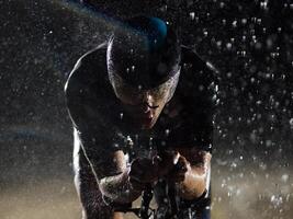 A triathlete braving the rain as he cycles through the night, preparing himself for the upcoming marathon. The blurred raindrops in the foreground and the dark, moody atmosphere in the background add photo