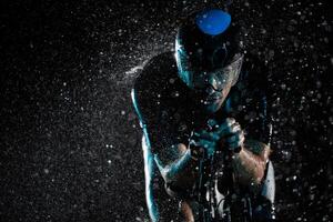 A triathlete braving the rain as he cycles through the night, preparing himself for the upcoming marathon. The blurred raindrops in the foreground and the dark, moody atmosphere in the background add photo