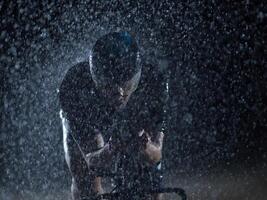 A triathlete braving the rain as he cycles through the night, preparing himself for the upcoming marathon. The blurred raindrops in the foreground and the dark, moody atmosphere in the background add photo