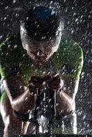 A triathlete braving the rain as he cycles through the night, preparing himself for the upcoming marathon. The blurred raindrops in the foreground and the dark, moody atmosphere in the background add photo