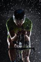 A triathlete braving the rain as he cycles through the night, preparing himself for the upcoming marathon. The blurred raindrops in the foreground and the dark, moody atmosphere in the background add photo