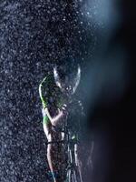 A triathlete braving the rain as he cycles through the night, preparing himself for the upcoming marathon. The blurred raindrops in the foreground and the dark, moody atmosphere in the background add photo