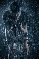 A triathlete braving the rain as he cycles through the night, preparing himself for the upcoming marathon. The blurred raindrops in the foreground and the dark, moody atmosphere in the background add photo