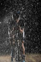 A triathlete braving the rain as he cycles through the night, preparing himself for the upcoming marathon. The blurred raindrops in the foreground and the dark, moody atmosphere in the background add photo